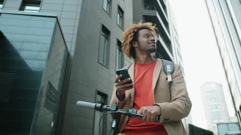 Hombre-Afroamericano-De-Pie-Con-E-scooter-En-La-Ciudad-Y-Usando-Un-Teléfono-Inteligente